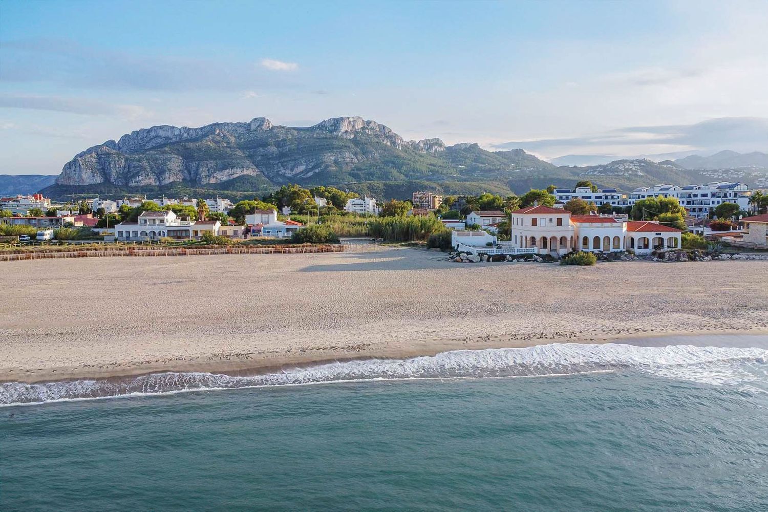Luftaufnahme des Sandstrandes mit sanften Wellen und einem klassischen Küstenhaus mit Bögen, im Hintergrund das Montgó-Massiv.