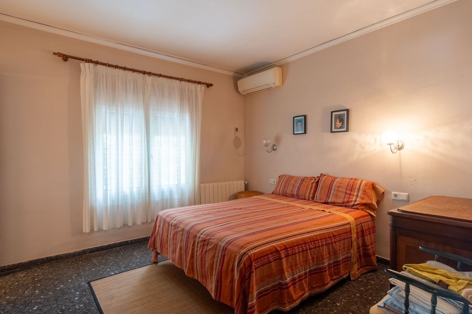 Schlafzimmer mit Doppelbett, gestreifter Bettwäsche, Fenster mit hellen Vorhängen, dunklem Boden und Klimaanlage.