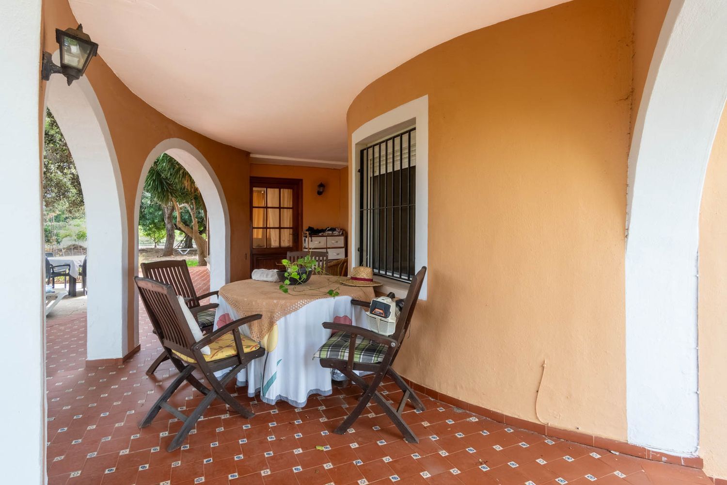 Überdachte Terrasse mit rotbraunen Fliesen und ockerfarbener Rundwand. Weiße Bögen führen zum Garten, ausgestattet mit einem runden Tisch und Holzstühlen.