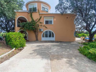 Außenansicht der pfirsichfarbenen Villa mit abgerundetem Gebäudeteil, Balkon und Haupteingang. Eine große Betonzufahrt und ein alter Garten mit Pool sind im Hintergrund sichtbar.