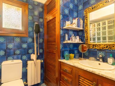 Badezimmer mit charakteristischen blauen Musterfliesen, einem Holzwaschtisch mit weißer Marmorplatte und einem verzierten Goldspiegel. Ein mattiertes Holzfenster spendet Tageslicht.