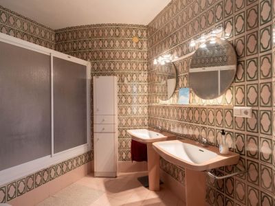 Badezimmer mit markanten grün-beigen Wandfliesen, Doppelwaschtisch mit zwei Waschbecken, runden Spiegeln mit Beleuchtung, Duschkabine und hohem Spiegelschrank.