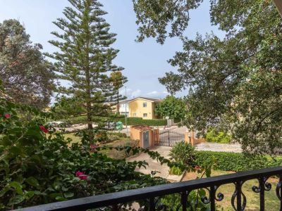 Balkonblick mit schmiedeeisernem Geländer und üppiger mediterraner Vegetation, die auf einen Zufahrtsweg und ein gelbes Haus im Hintergrund unter klarem Himmel blickt.