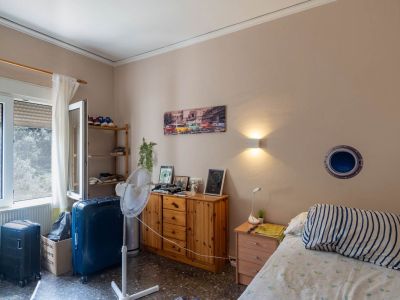 Funktionales Schlafzimmer mit Einzelbett, Nachttisch und Kommode aus Holz, beleuchtet durch Tageslicht vom Fenster und eine Wandleuchte. Dunkler, gesprenkelter Boden.