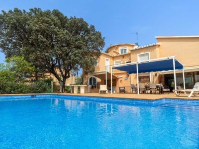 Großer blau gefliester Pool mit angrenzender, weitläufiger Terrasse und Essbereich im Freien unter einer blauen Pergola. Die ockerfarbene Hausfassade wird von einem alten Baum eingerahmt.