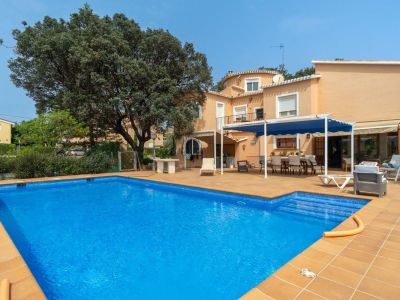 Großer rechteckiger Swimmingpool mit Terrakotta-Fliesenrand und weitläufiger Terrasse. Ein Essbereich im Freien unter einer blauen Pergola und die pfirsichfarbene Hausfassade sind zu sehen.