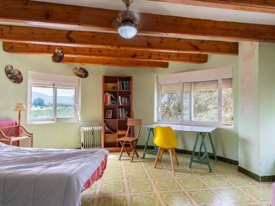 Helles Schlafzimmer mit sichtbaren Holzbalken und hellgrünen Wänden. Es verfügt über gemusterte Bodenfliesen, Fenster mit Naturblick und einen Arbeitsbereich.