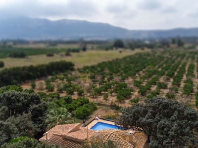 Luftaufnahme des Hauses mit Ziegeldach und blauem Pool, umgeben von mediterraner Vegetation, vor weiten Feldern und fernen Bergen.