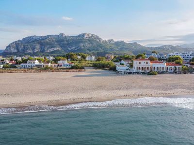 Luftaufnahme des Sandstrandes mit sanften Wellen und einem klassischen Küstenhaus mit Bögen, im Hintergrund das Montgó-Massiv.