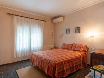 Schlafzimmer mit Doppelbett, gestreifter Bettwäsche, Fenster mit hellen Vorhängen, dunklem Boden und Klimaanlage.