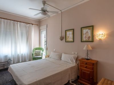 Schlafzimmer mit Doppelbett, Nachttisch aus Holz und Lampe. Es verfügt über einen Deckenventilator, einen grünen Korbstuhl und einen verglasten Zugang nach draußen.