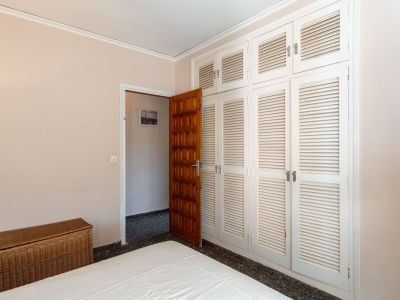 Schlafzimmer mit einem geräumigen Einbauschrank mit weißen Lamellentüren, einer dunklen Holztür und einem Bett. Eine Flechtkommode ergänzt den Raum.