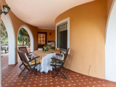 Überdachte Terrasse mit rotbraunen Fliesen und ockerfarbener Rundwand. Weiße Bögen führen zum Garten, ausgestattet mit einem runden Tisch und Holzstühlen.