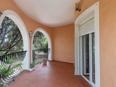 Überdachte Terrasse mit Terrakottafliesen, eleganten Bögen und schmiedeeisernen Geländern. Bietet Blick auf Bäume und Zugang durch eine Schiebetür.