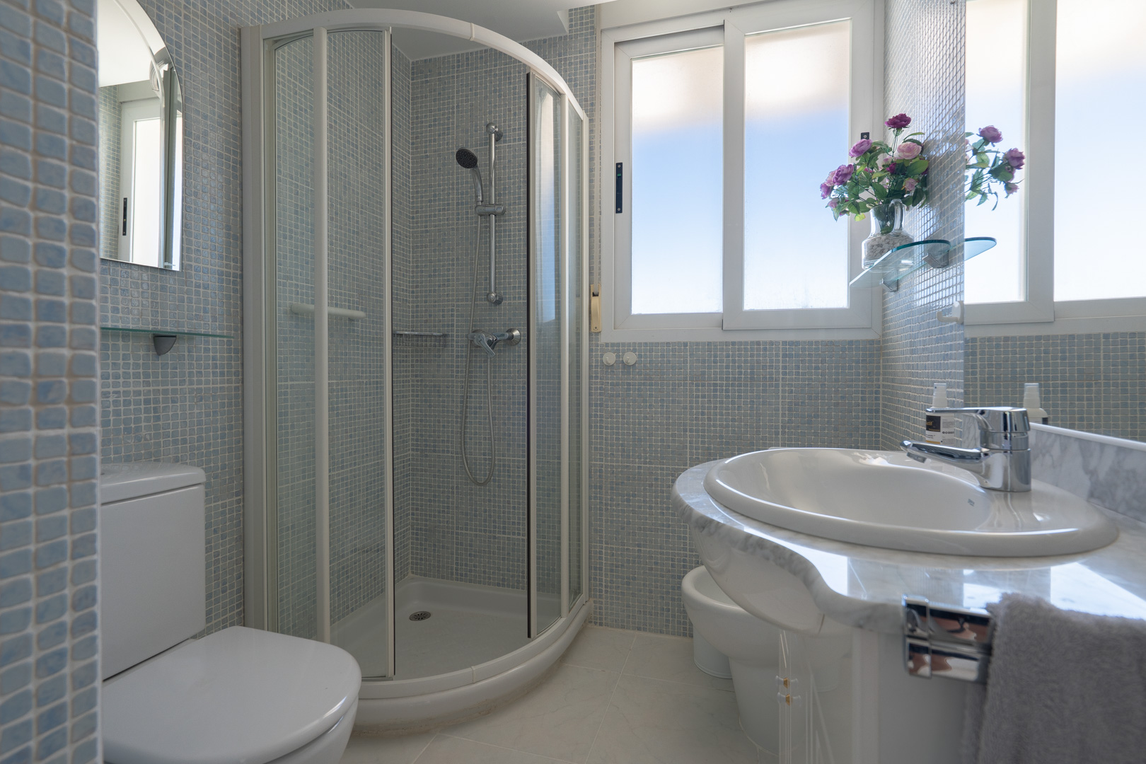 Badezimmer mit Eckdusche, WC, Bidet und Waschtisch mit Marmoroptik. Hellblaue Mosaikfliesen an den Wänden und Fenster für natürliches Licht.