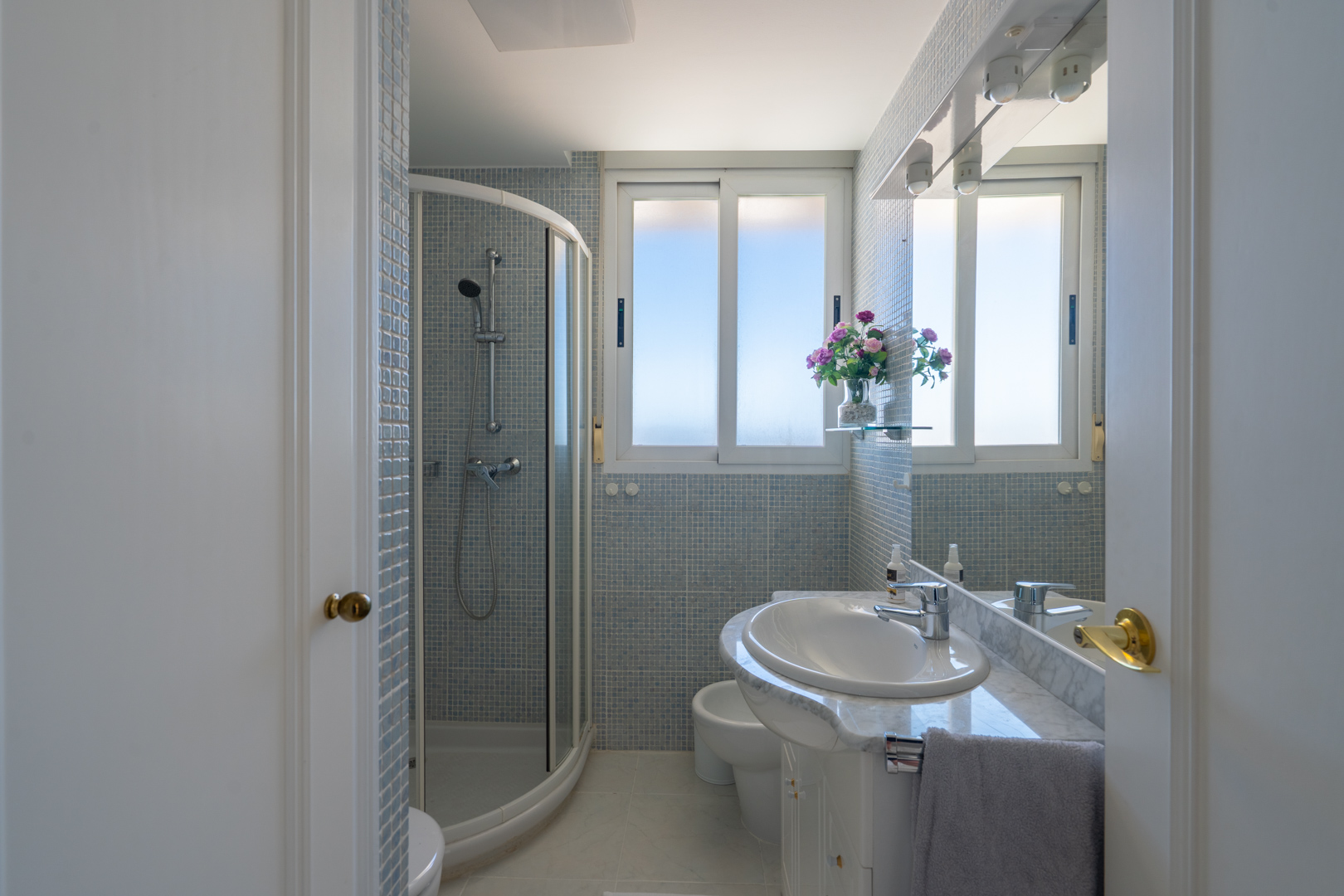 Badezimmer mit Eckduschkabine, weißem Waschtisch mit Marmorplatte und hellblauen Mosaikfliesen. Fenster für Tageslicht und beleuchteter Spiegel.