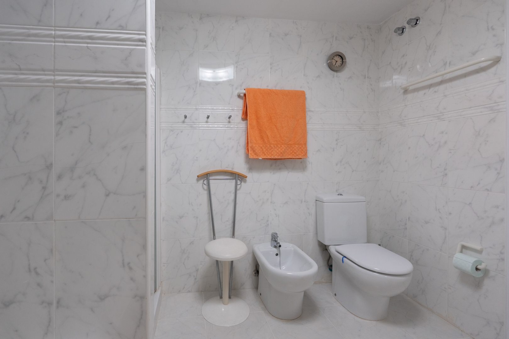 Badezimmer mit weißen Marmor-Effekt-Fliesen, ausgestattet mit WC, Bidet und einem praktischen Duschbereich mit Haltegriff. Eine Wanduhr und Handtuchhaken sind ebenfalls vorhanden.