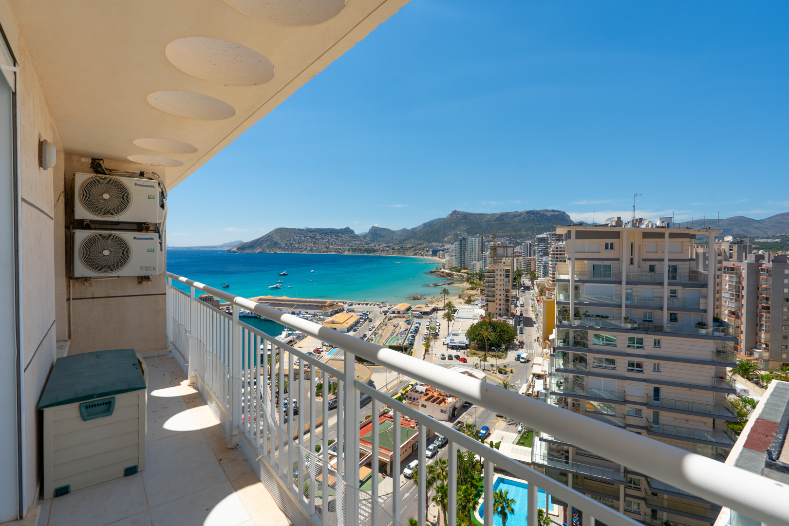 Balkon mit Panoramablick auf Calpes Yachthafen, die türkisfarbene Bucht und die Stadtküste, eingerahmt von einem weißen Geländer und externen Klimaanlagen.