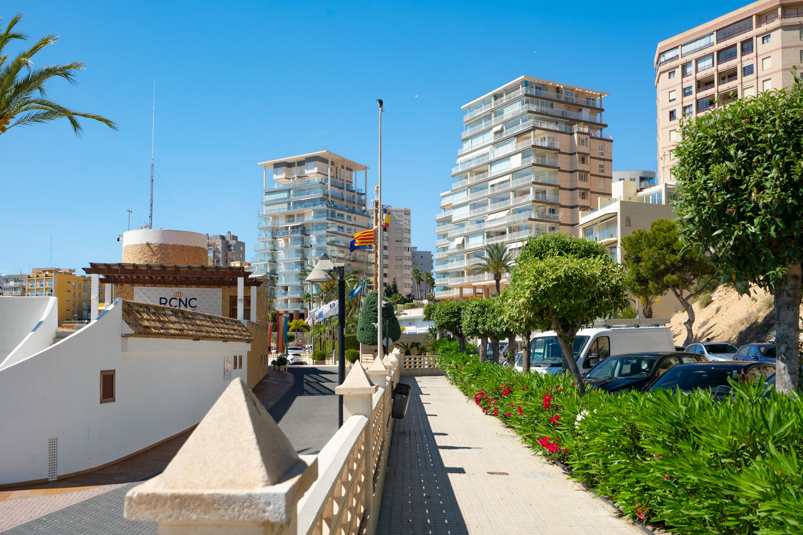 Blick auf die belebte Promenade von Calpe, mit modernen Hochhauswohnungen und dem Gebäude des Königlichen Yachtclubs unter klarem blauem Himmel.