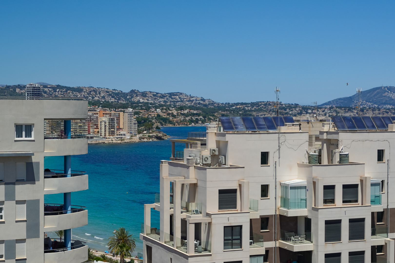 Blick von einer oberen Etage auf moderne Küstenarchitektur, mit verglasten Balkonen und Solarpaneelen auf den Dächern, umrahmt vom türkisfarbenen Meer und fernen Hügeln.