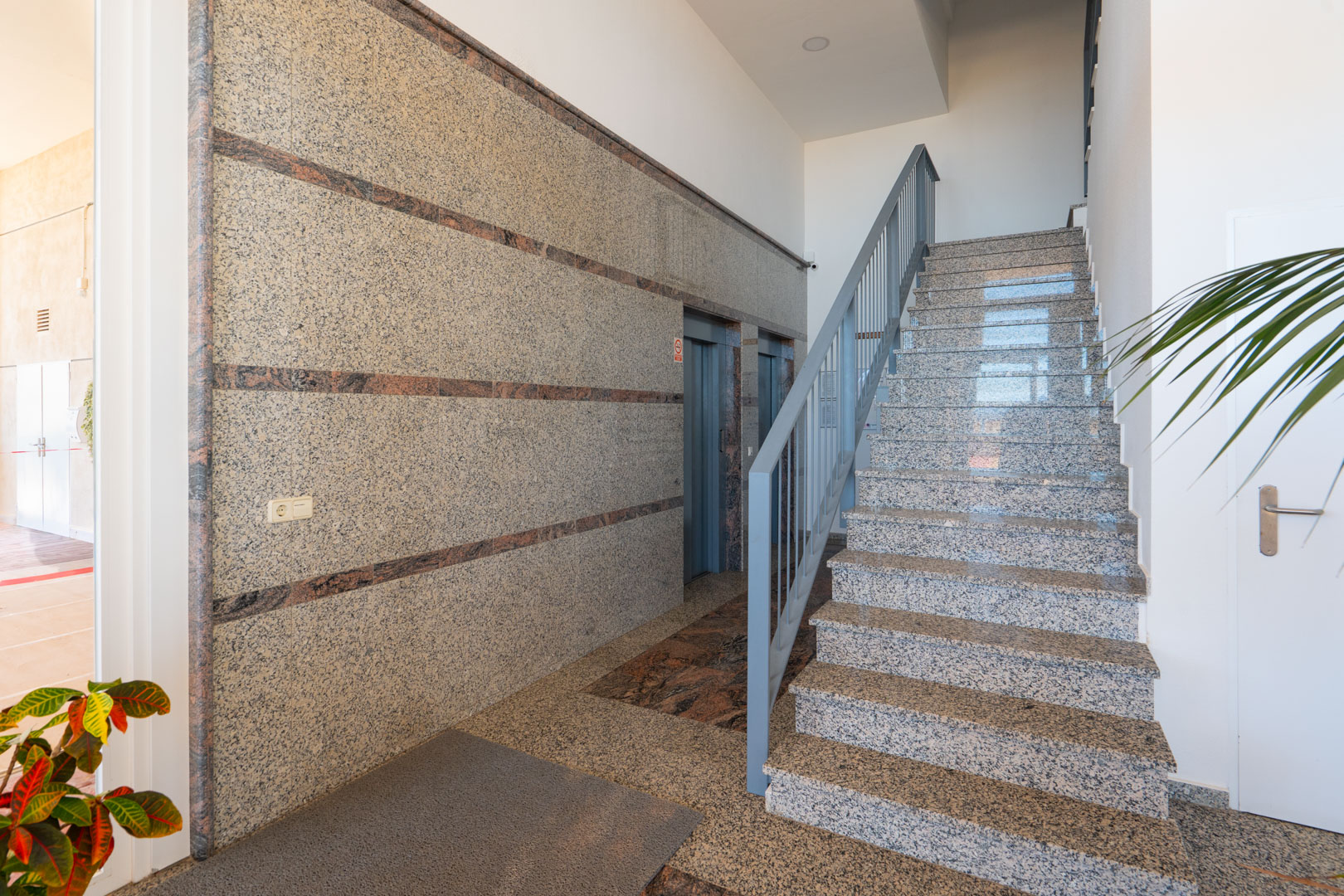 Der Gemeinschaftsbereich des Gebäudes zeigt polierte Granitwände mit dunklen Akzentstreifen, eine passende Granittreppe mit grauem Geländer und zwei Aufzüge, beleuchtet durch Einbaustrahler.