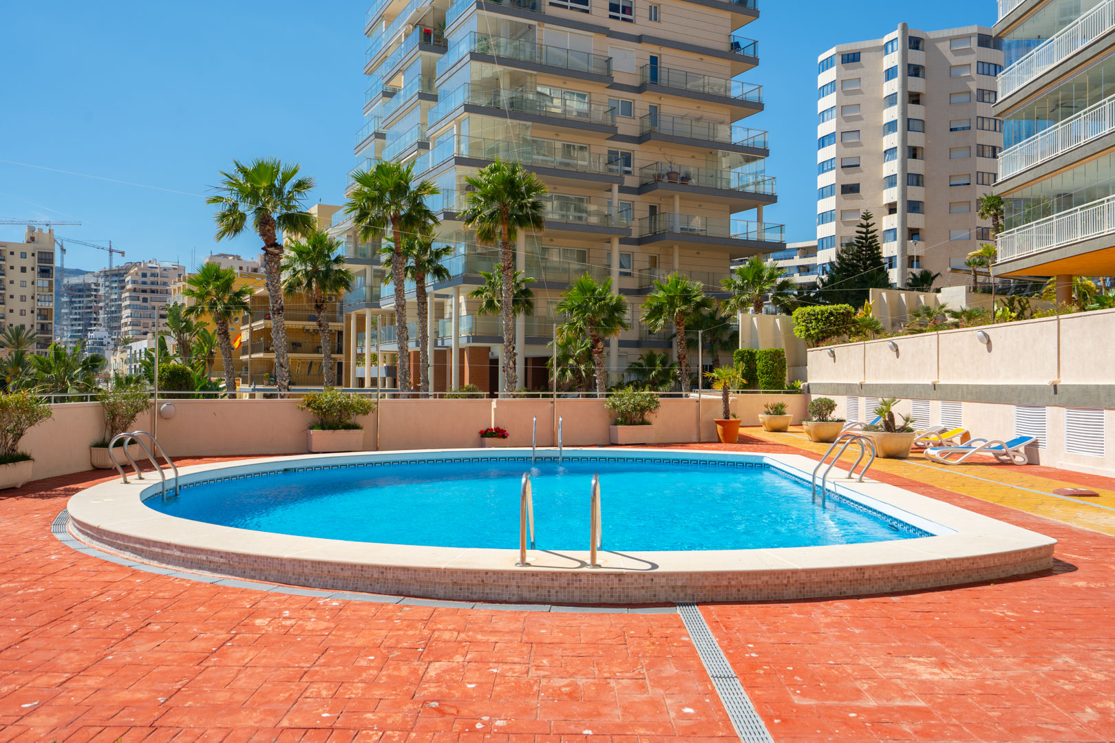Der Gemeinschaftspool mit blauem Wasser und Edelstahlstufen ist von einer Terrasse mit Terrakottafliesen und Palmen umgeben, dahinter moderne Wohngebäude.