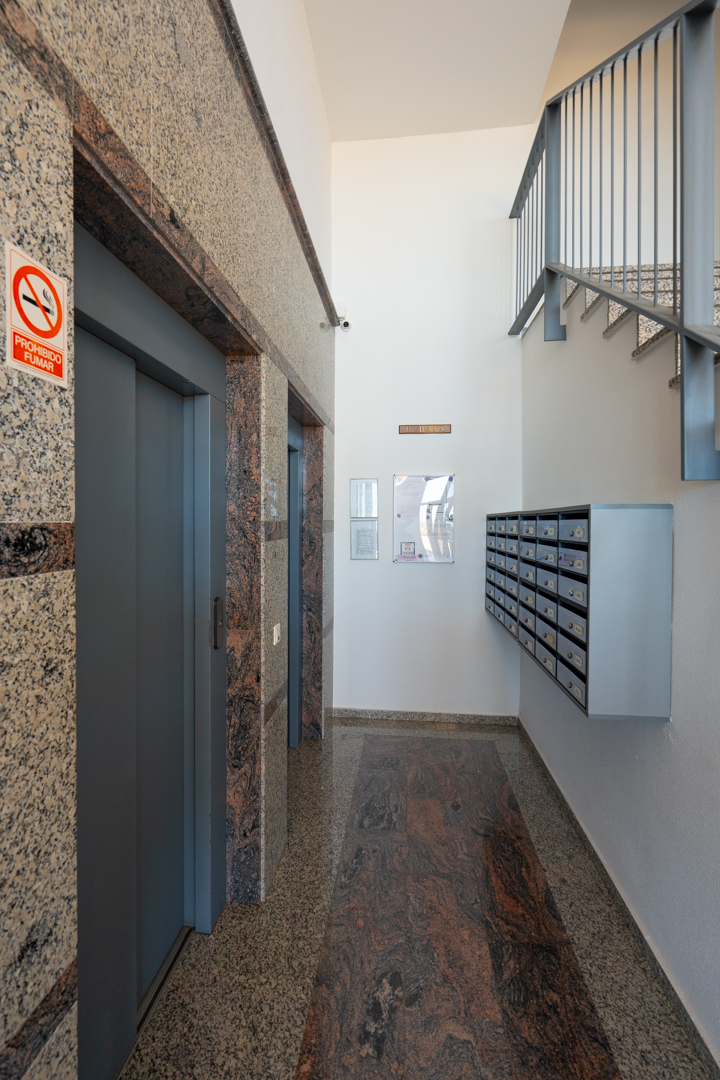 Eingangsbereich mit zwei Aufzugstüren, umrahmt von gesprenkeltem Granit, einem dunklen Granitbodenläufer, Briefkästen und einer Metalltreppe.