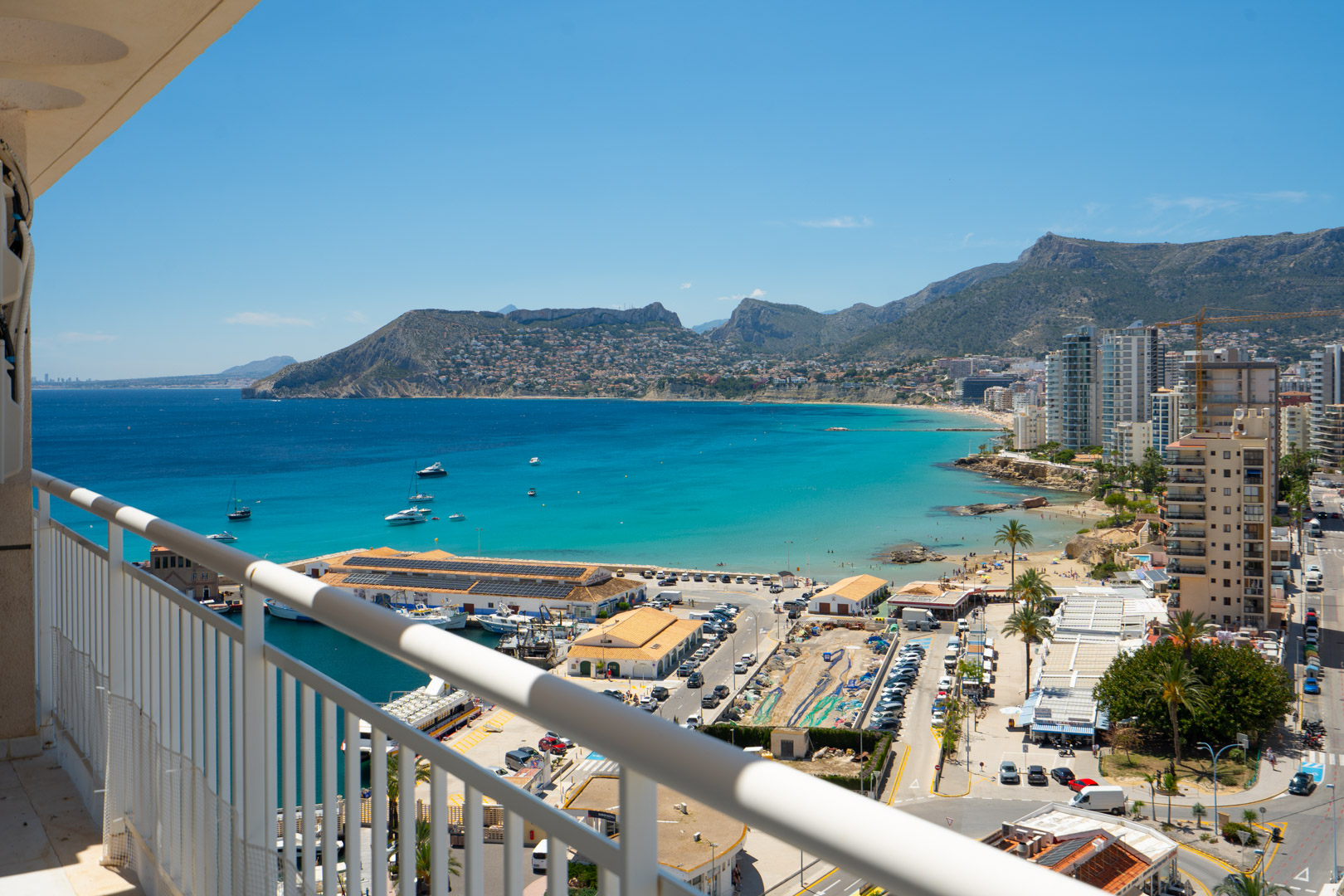 Erhöhter Terrassenblick über Calpes lebhaften Hafen, die türkisfarbene Bucht und die ferne Berglandschaft der Costa Blanca bei klarem Himmel.