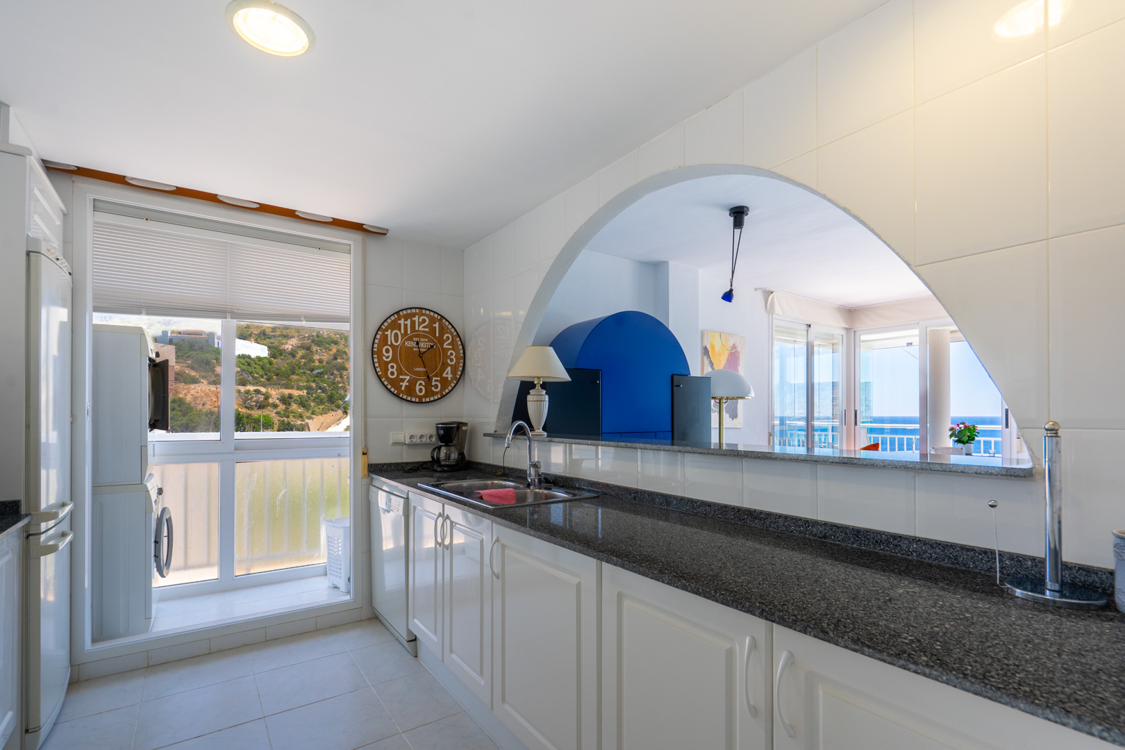 Funktionale Küche mit weißen Schränken, dunkler Granitarbeitsplatte und Waschküche am Fenster mit Blick auf den Hügel. Ein Bogen verbindet sie mit dem Wohnbereich und Meerblick.