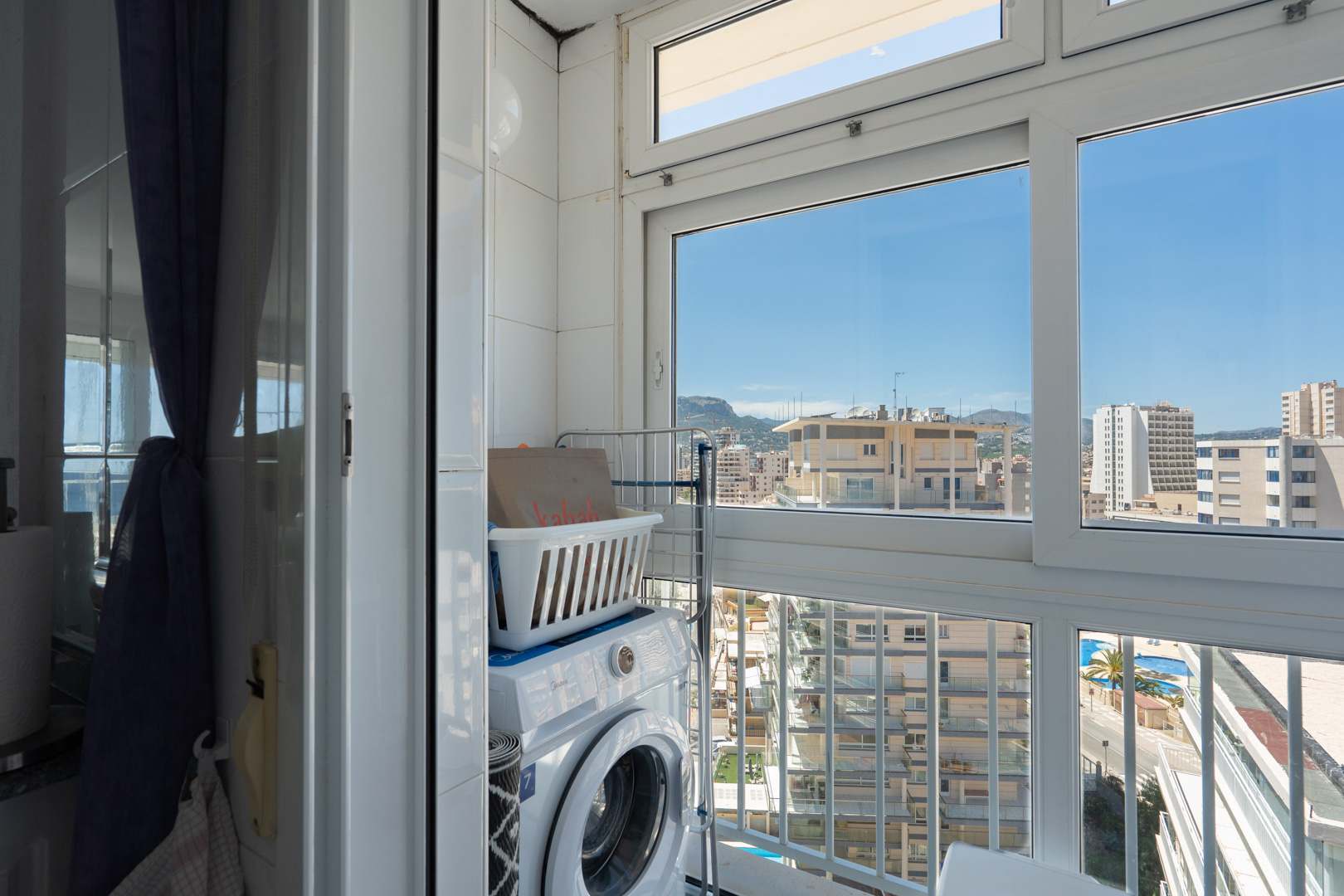 Funktionale Waschecke mit Waschmaschine und Wäschekorb, umgeben von Fliesenwänden und großen Fenstern, die einen urbanen Blick auf Gebäude, ferne Berge und einen Pool bieten.