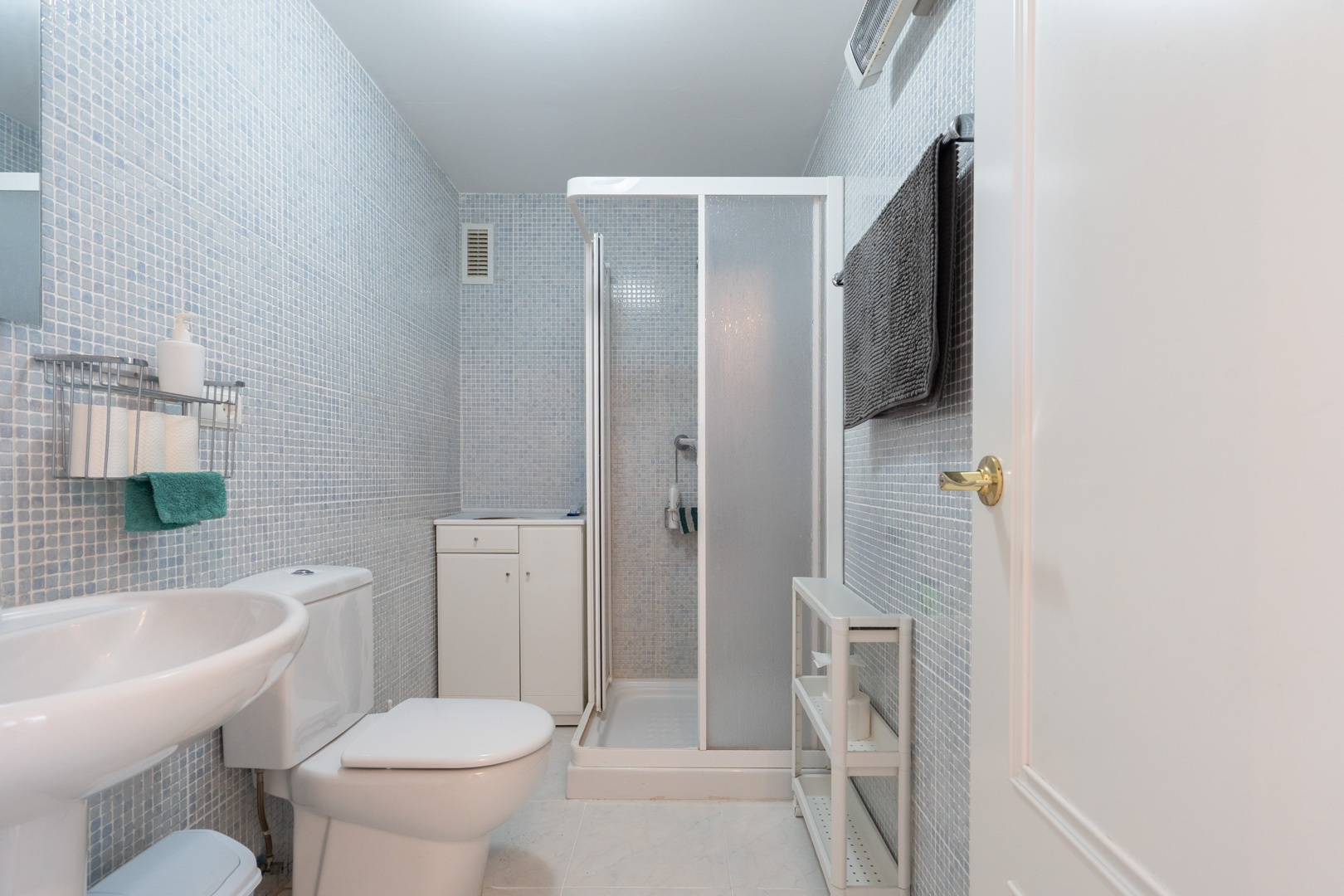 Funktionales Badezimmer mit hellblauen Mosaikfliesen an den Wänden, ausgestattet mit WC, Standwaschbecken und Duschkabine. Ein kleiner Unterschrank und zusätzliche Regale bieten Stauraum.