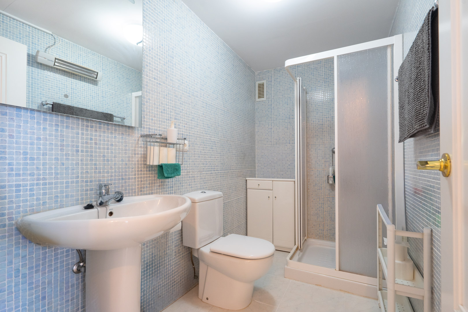 Funktionales Badezimmer mit hellblauen Mosaikfliesen, Standwaschbecken, WC, Duschkabine mit Milchglas und einem weißen Beistellschrank für Stauraum.