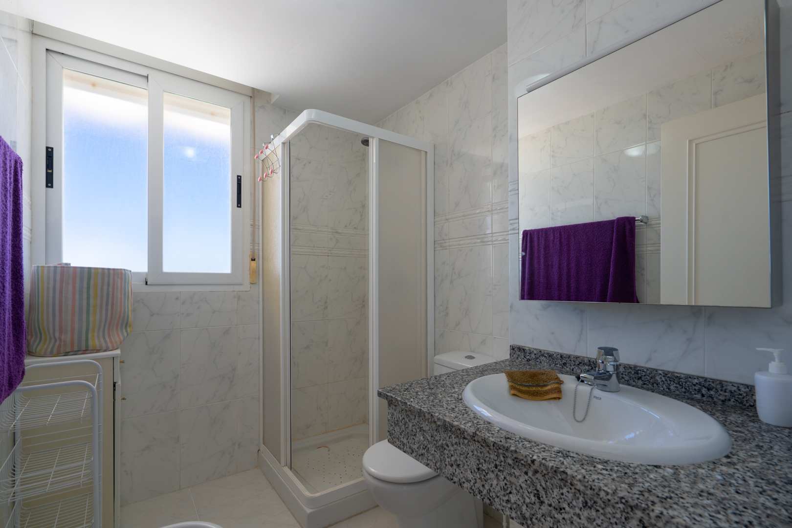 Funktionales Badezimmer mit weißen Marmor-Optik Fliesen, einem Waschtisch aus gesprenkeltem Granit mit ovalem Waschbecken, einer Eckdusche und Tageslichtfenster.