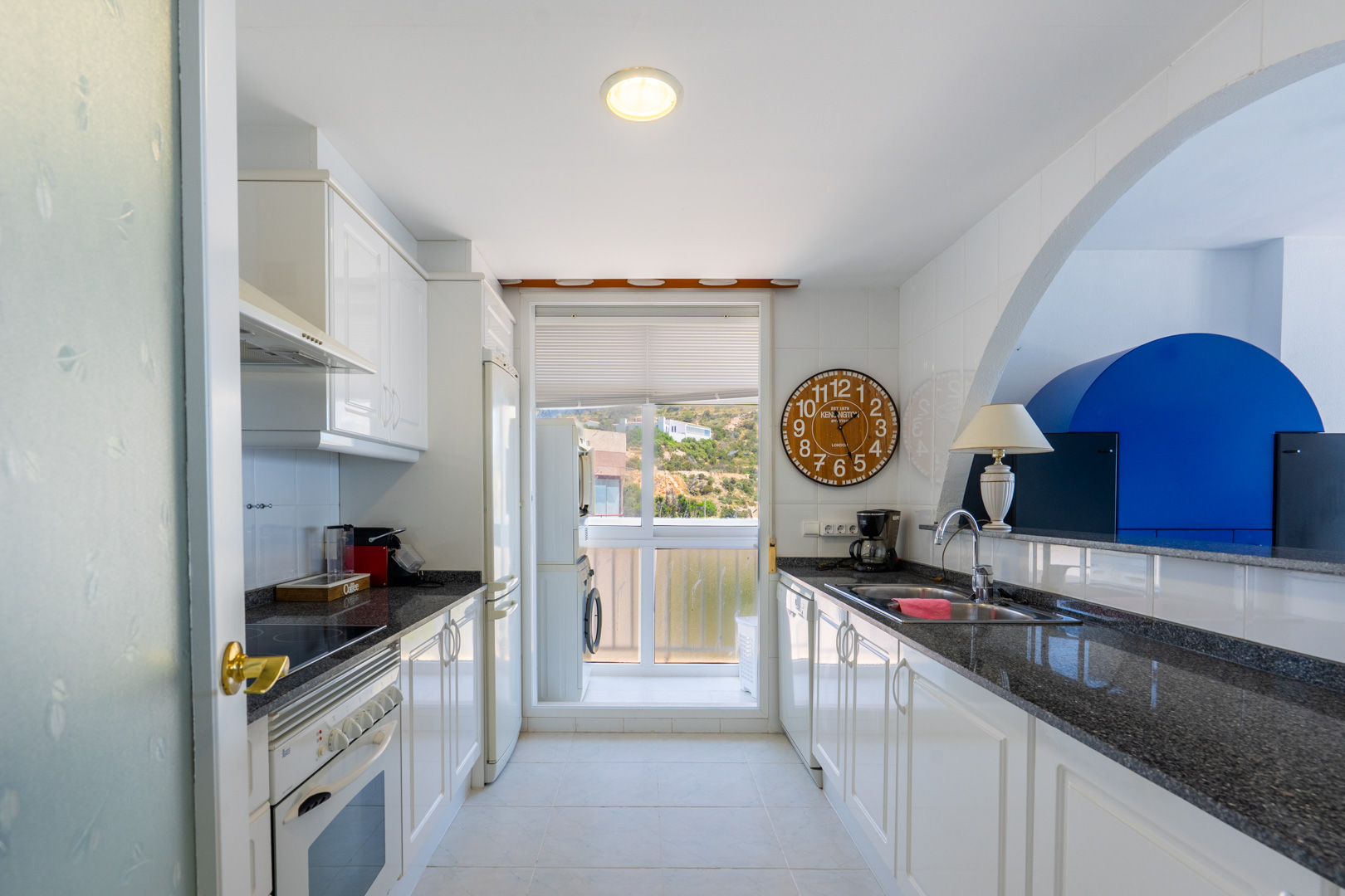 Galeerenküche mit weißen Schränken, dunklen Granitarbeitsplatten und Fliesenrückwand. Backofen, Kühlschrank und Waschmaschine im angrenzenden Hauswirtschaftsraum, mit Hügelblick.