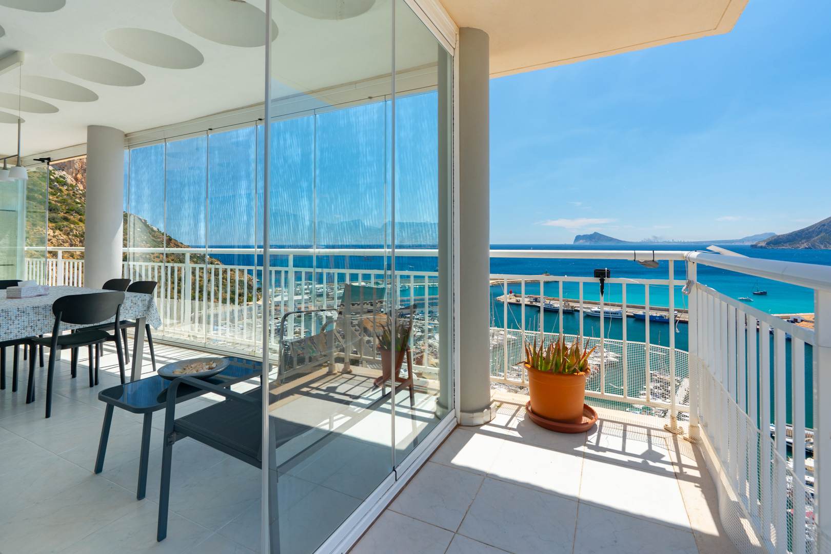 Geräumige Terrasse mit Glasschiebevorhängen, die einen Essbereich umschließen und direkten Blick auf den Yachthafen und die Bucht von Calpe bieten. Ein offener Bereich mit weißem Geländer und Pflanze ist sichtbar.