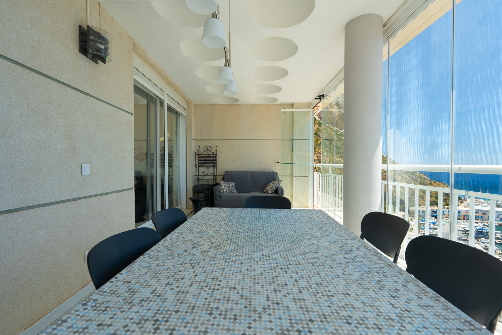 Geräumige überdachte Terrasse mit Mosaik-Esstisch, Sitzgelegenheiten und Glasvorhängen, die Panoramablicke auf den Hafen, das Meer und die Berge freigeben.