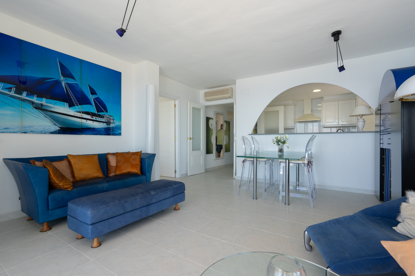Geräumiger Wohn-Essbereich mit blauem Sofa, Glastisch und transparenten Stühlen, akzentuiert durch ein großes Segelboot-Kunstwerk. Ein Bogen führt zur modernen, halboffenen Küche.