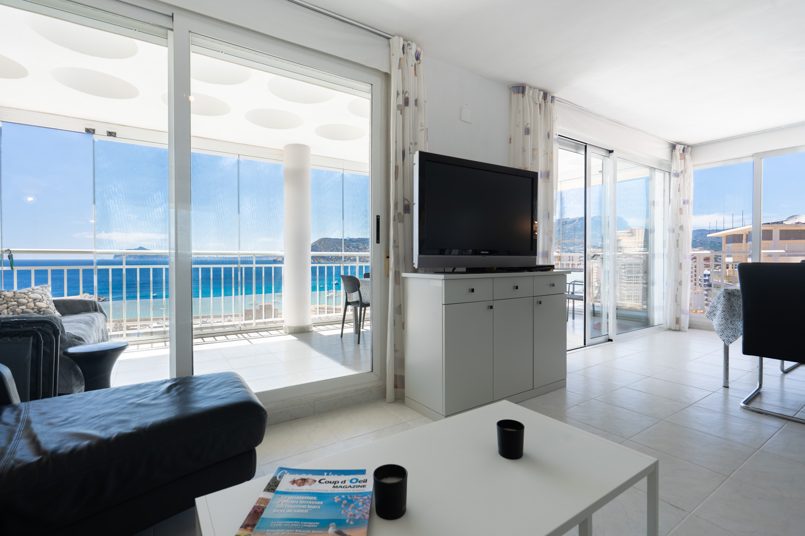 Geräumiger Wohnbereich mit Fliesenboden, TV-Möbel und Sofa, der sich zu einer verglasten Terrasse mit Panoramameerblick und gemusterter Decke öffnet.