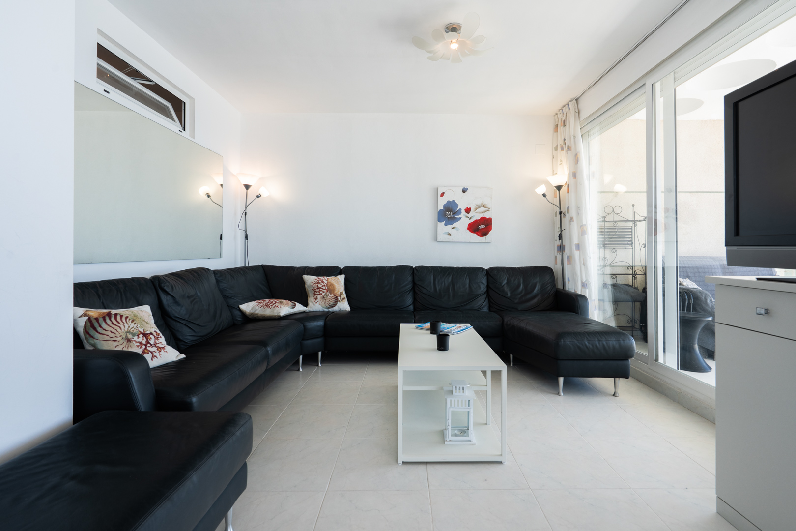 Geräumiges Wohnzimmer mit großem schwarzen Ecksofa und Zierkissen, weißen Fliesenboden und modernem weißen Couchtisch. Glasschiebetüren führen direkt zur Terrasse.