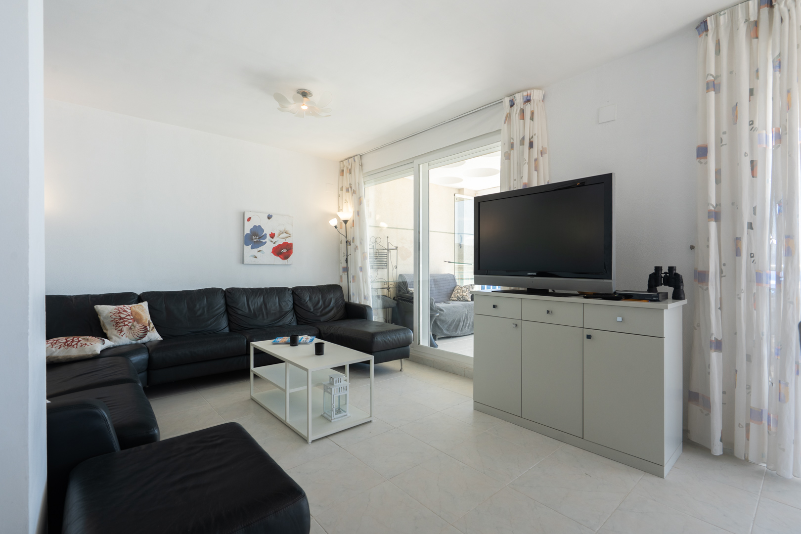Geräumiges Wohnzimmer mit schwarzem Lederecksofa, weißem Couchtisch und Fliesenboden. Glasschiebetüren führen zu einer Terrasse, ergänzt durch einen TV-Schrank und gemusterte Vorhänge.
