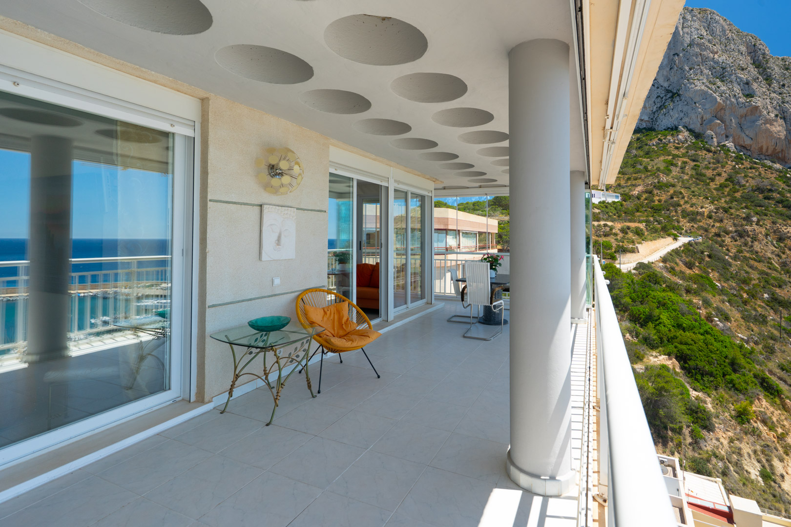 Große überdachte Terrasse mit markanten kreisförmigen Deckenmustern, Glasschiebetüren, eleganten Fliesenböden und doppeltem Blick auf das Meer und den Peñón de Ifach.
