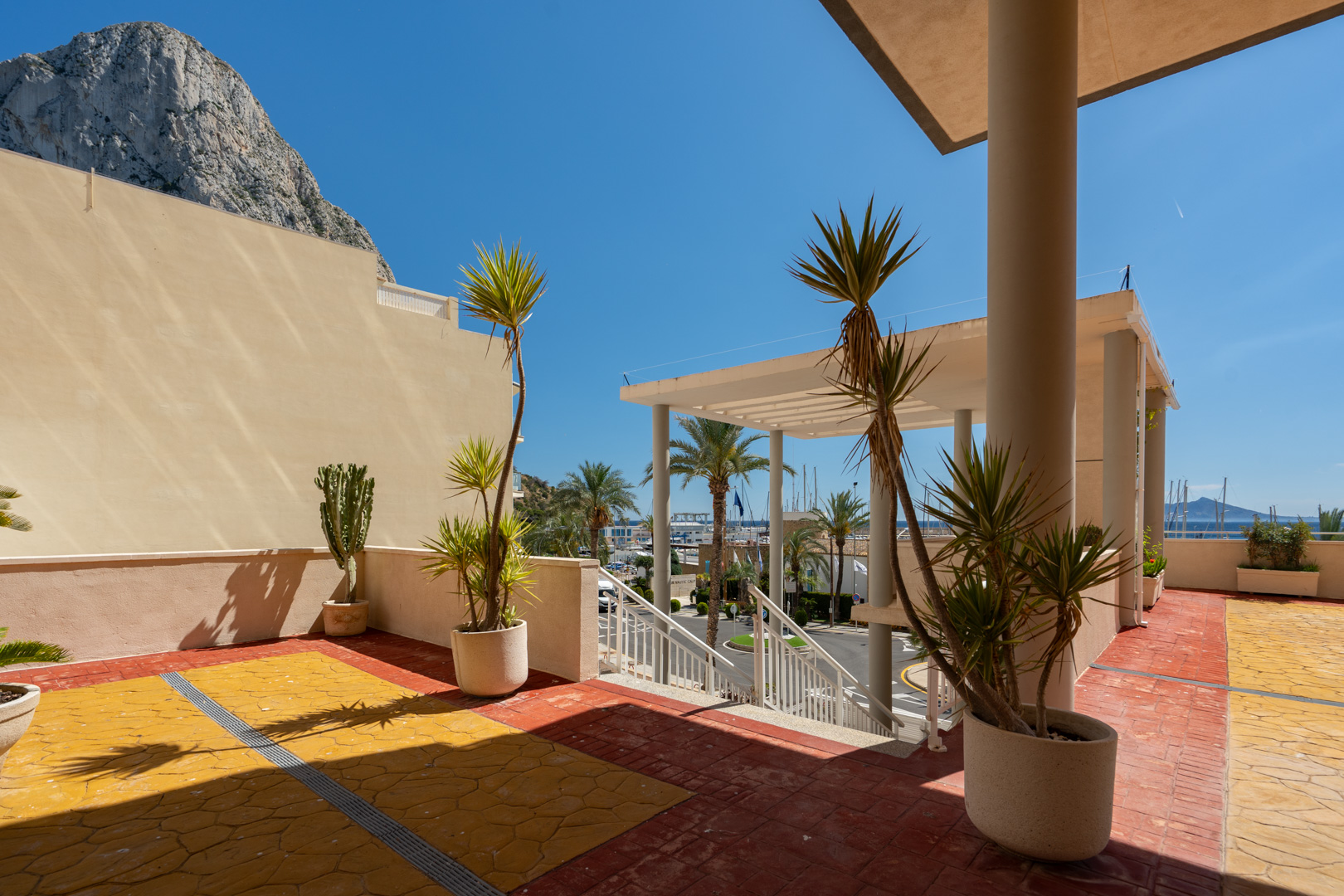 Großzügige Außenterrasse mit rot-gelben Fliesen, mediterranen Pflanzen und direktem Blick auf den Hafen von Calpe und den Peñón de Ifach unter blauem Himmel.