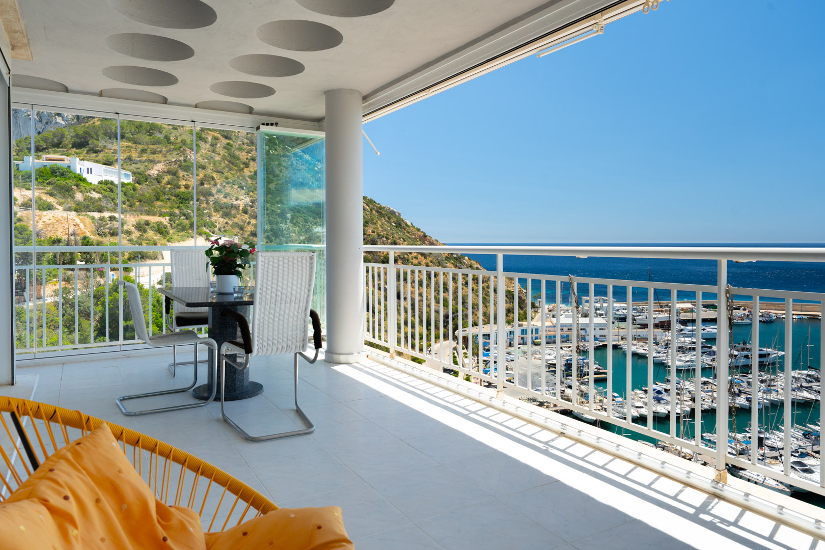 Großzügige geflieste Terrasse mit Esstisch und Glasvorhängen, die einen direkten Panoramablick auf den Hafen von Calpe, das Mittelmeer und grüne Hügel bietet.