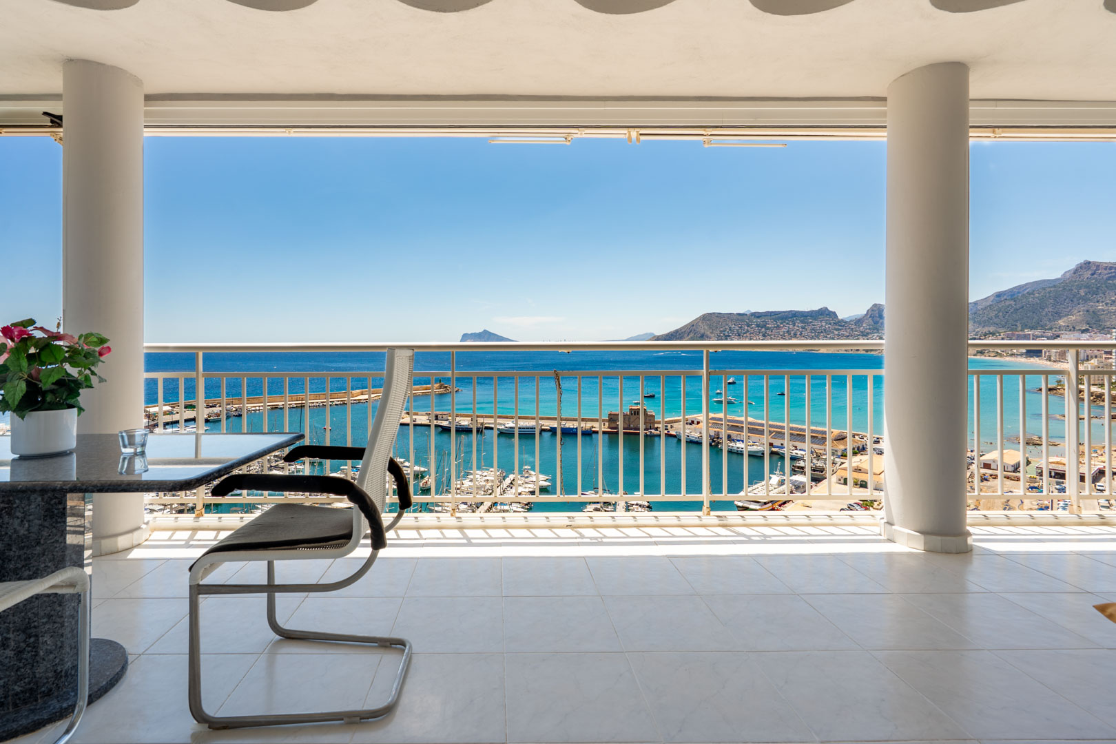 Großzügige überdachte Terrasse mit weißen Fliesen, die einen direkten Blick auf den Hafen von Calpe und das Mittelmeer bei strahlend blauem Himmel bietet.
