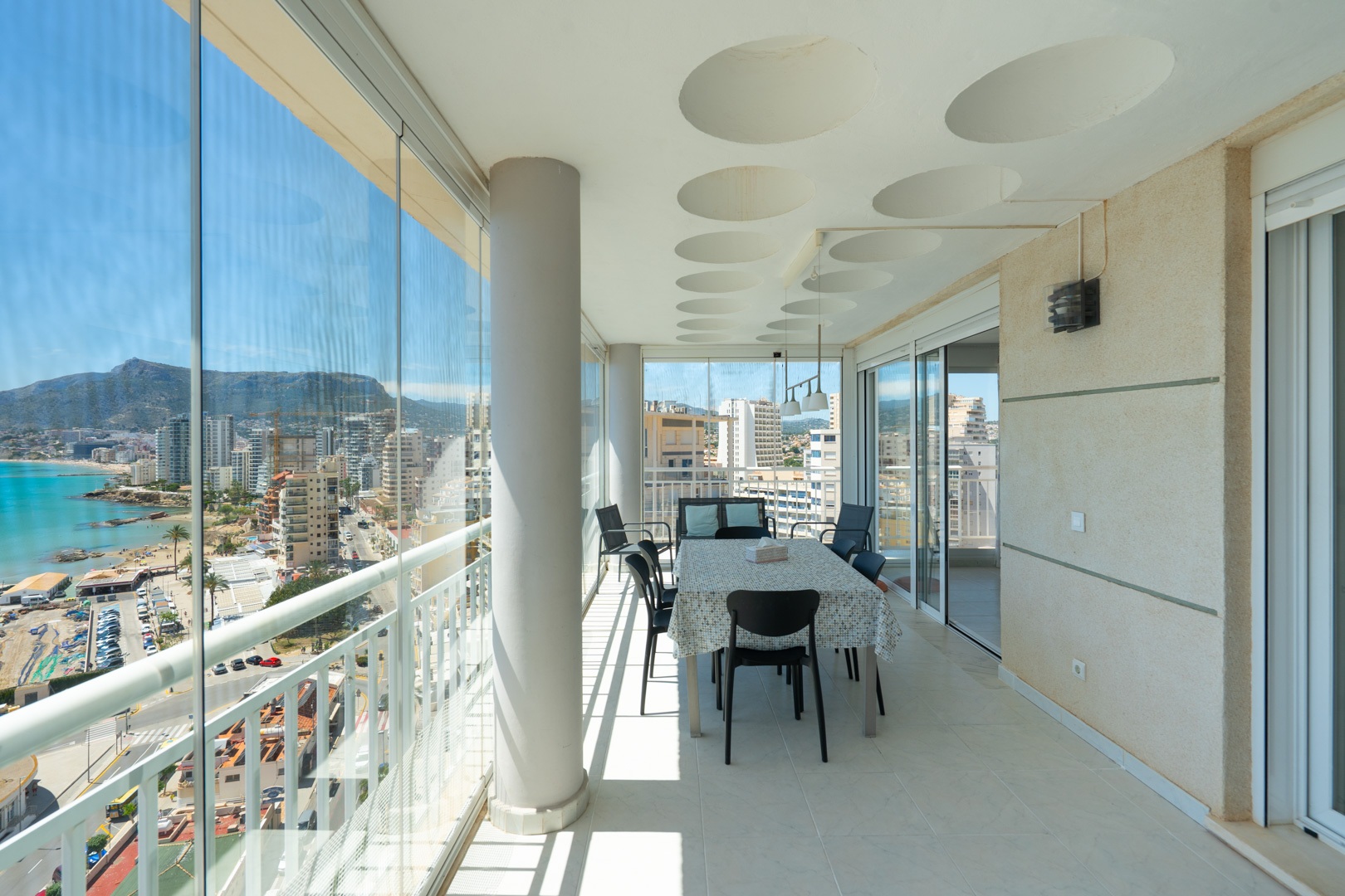 Großzügige verglaste Terrasse mit Essbereich und Panoramablick auf Meer, Hafen und Berge von Calpe. Sie verfügt über Glasvorhänge, eine Decke mit runden Akzenten und einen gefliesten Boden.
