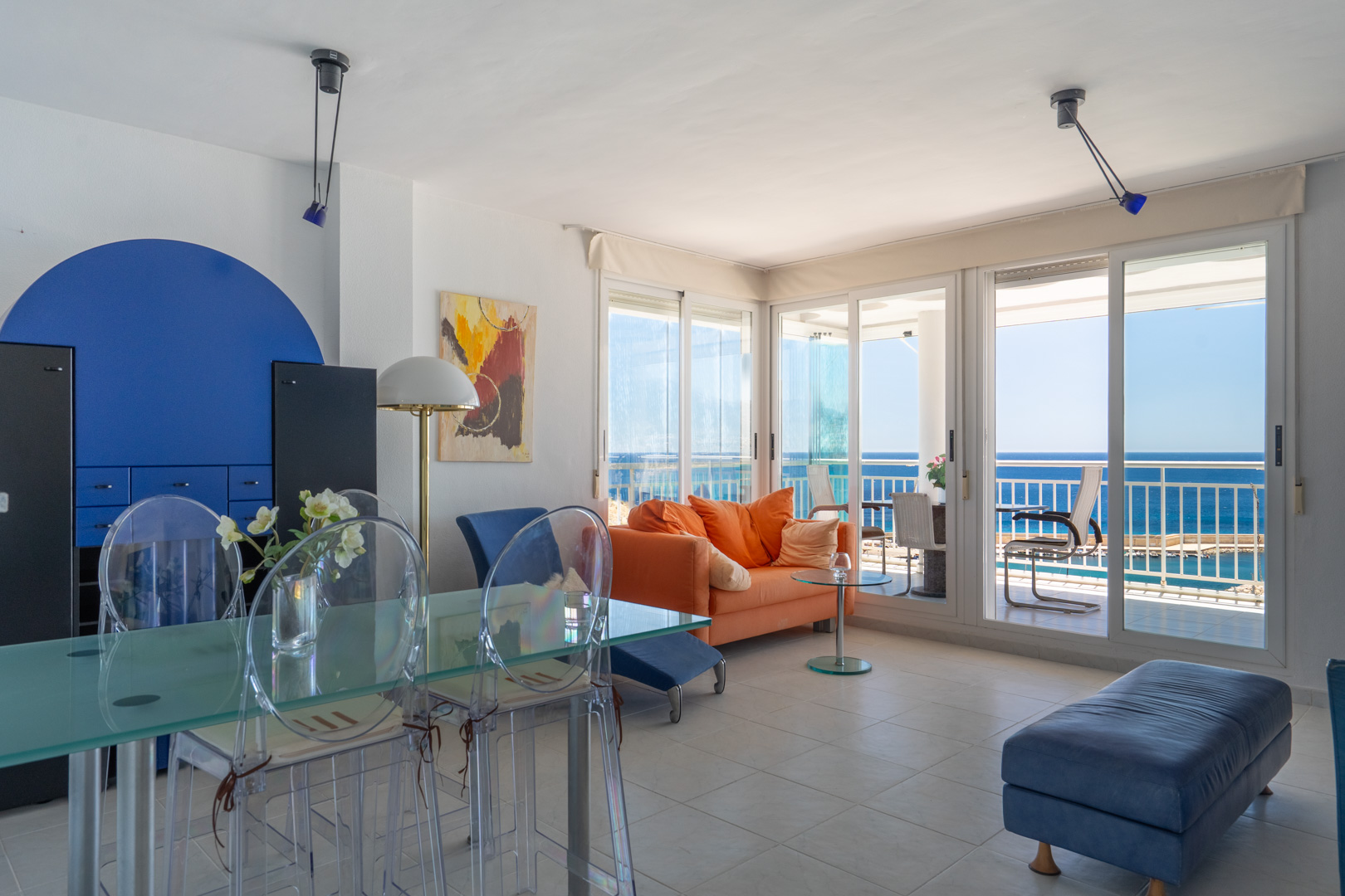 Großzügiger Wohn-Essbereich mit orangefarbenem Sofa, Glastisch und transparenten Stühlen, akzentuiert durch eine blaue Wand. Große Schiebetüren führen zu einer Terrasse mit direktem Meerblick.