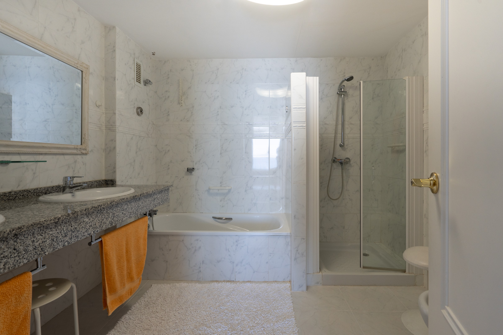 Hauptbadezimmer mit Doppelwaschtisch auf grauer Granitplatte, integrierter Badewanne und separater Duschkabine, komplett gefliest mit weißen Marmoreffekt-Fliesen.