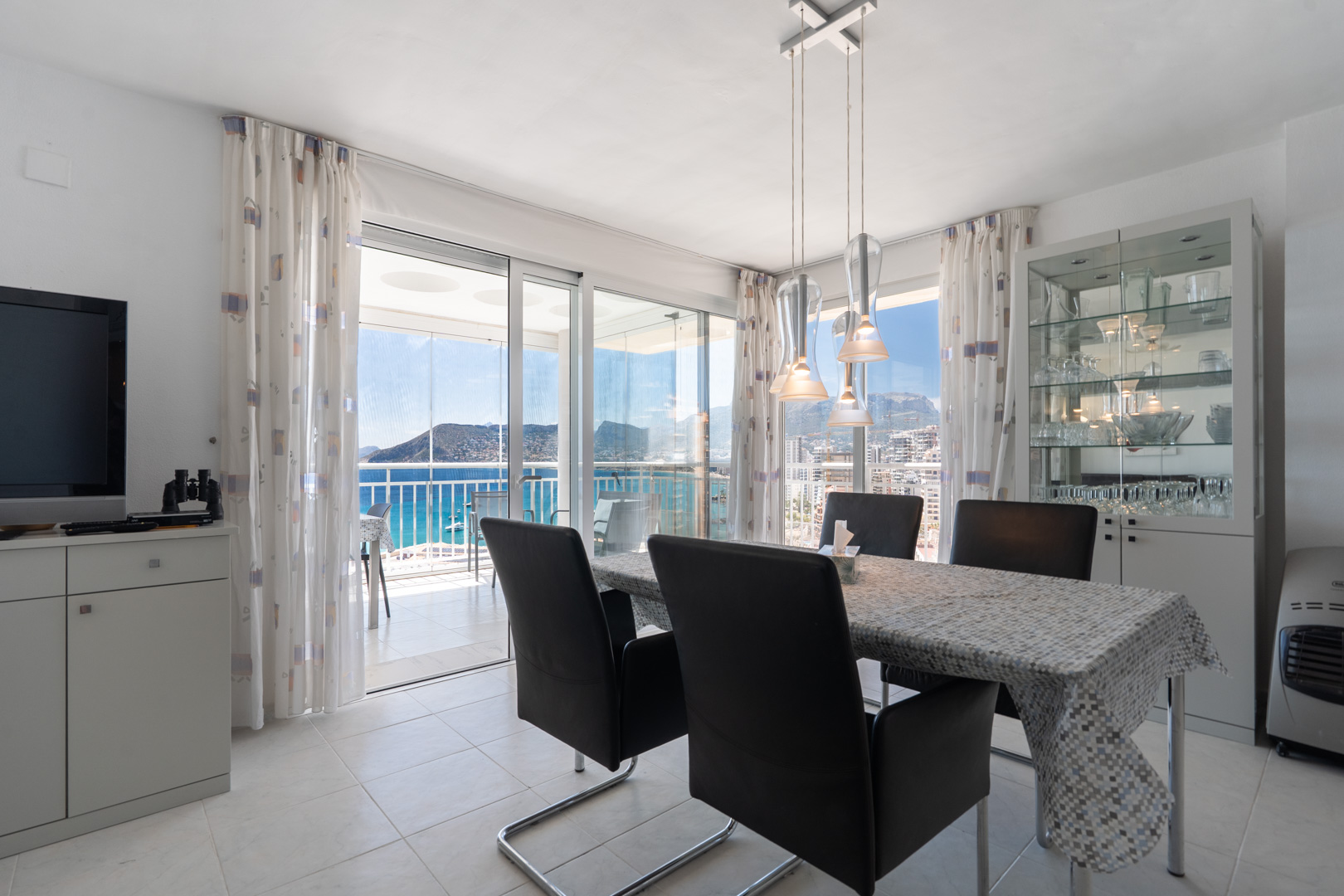 Heller Essbereich mit modernem Tisch und Stühlen, direkter Zugang zur verglasten Terrasse mit Panoramablick auf Meer und Peñón de Ifach. Vitrine mit Geschirr.