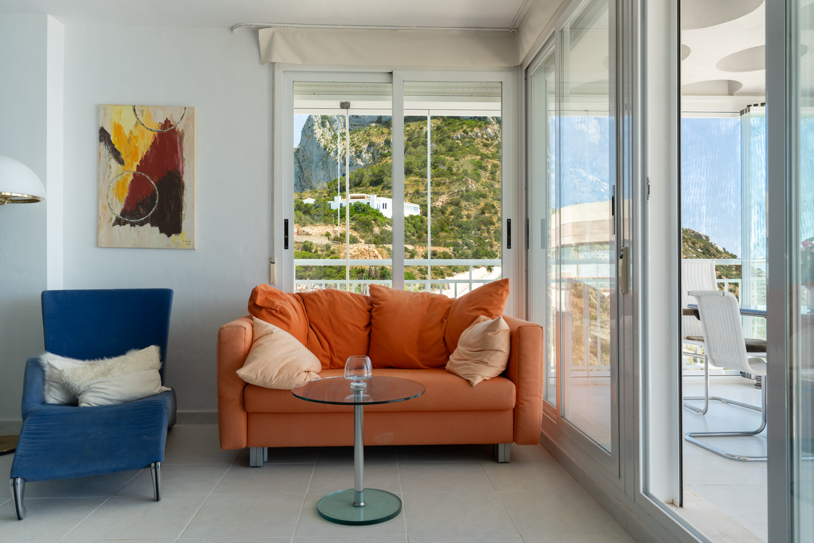 Heller Wohnbereich mit orangefarbenem Sofa und blauem Sessel. Große Schiebetüren führen zu einer Terrasse mit Bergblick, Außenmöbeln und einer Decke mit markantem Kreisdesign.