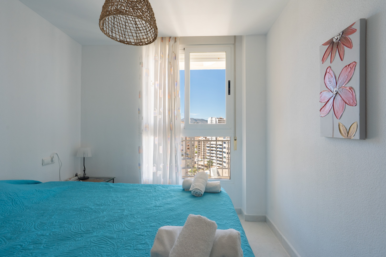 Helles Schlafzimmer mit blauem Steppbett und weißen Handtüchern. Das Fenster bietet Stadt- und Bergblick, ergänzt durch eine geflochtene Hängelampe und ein Blumenbild.